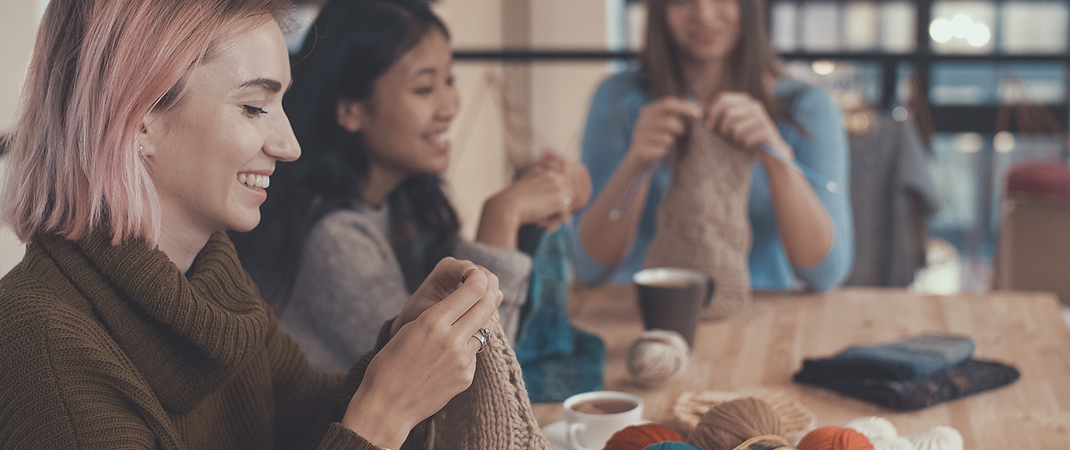 Strickende Hände mit Wolle, im Hintergrund Teilnehmerinnen bei einem Strick- und Häkeltreffen
